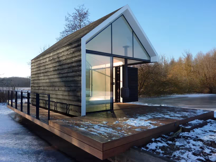 Casita de madera negra sobre su plataforma junto al lago helado, con barandilla y hastial de vidrio