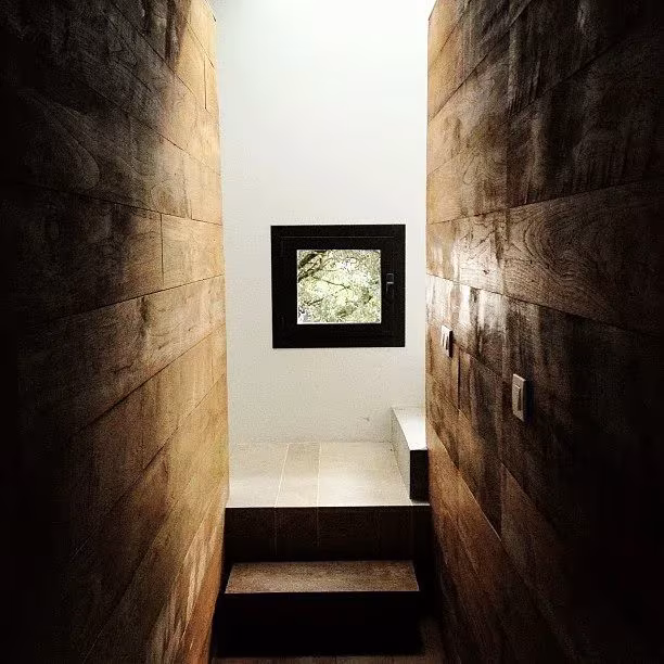 Paso estrecho entre muros de madera oscura con peldaños hacia una ventana cuadrada en la oficina de ÁBATON