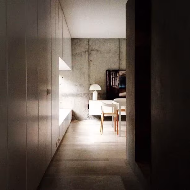Pasillo entre armarios grises y muros de hormigón que lleva a una sala con mesa blanca y lámpara
