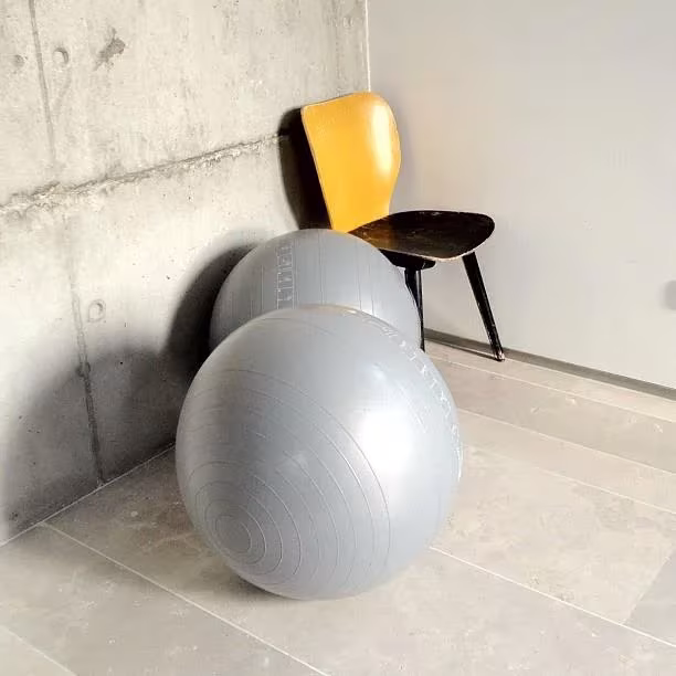 Silla de respaldo amarillo y dos pelotas de pilates grises en un rincón de hormigón de la oficina de ÁBATON