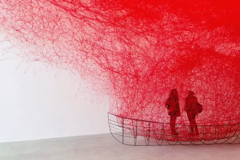 Instalación de hilos rojos de Chiharu Shiota que envuelve una estructura de barca, con dos visitantes dentro