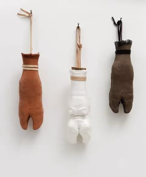 Tres piezas artesanales con forma de guante en cuero, cerámica blanca y tela, colgadas de una pared