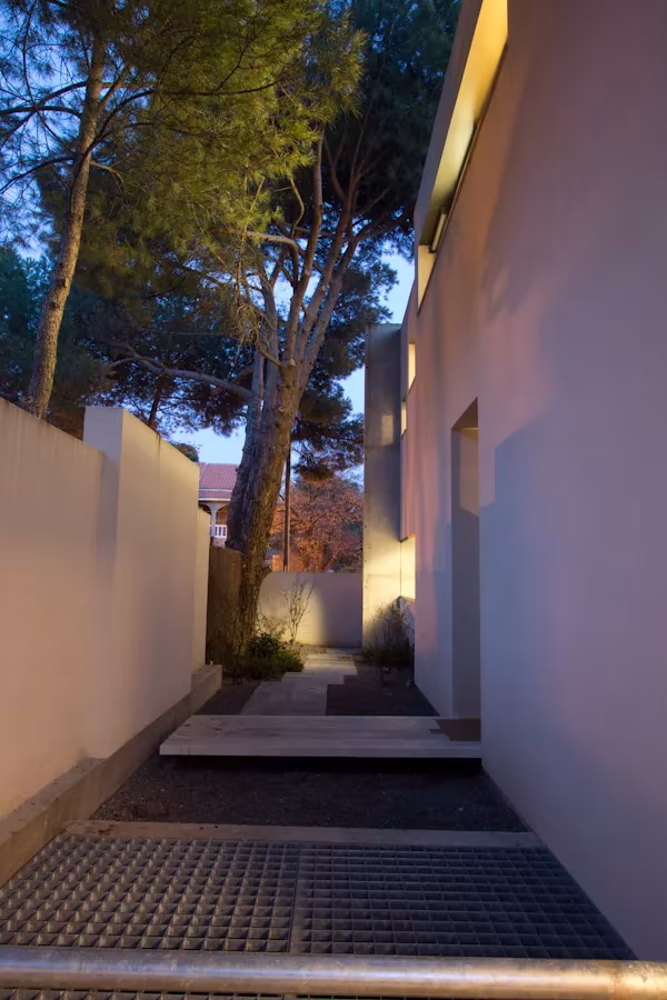 Pasillo exterior entre muros blancos de la oficina de ÁBATON al atardecer, con rejilla metálica y pinos