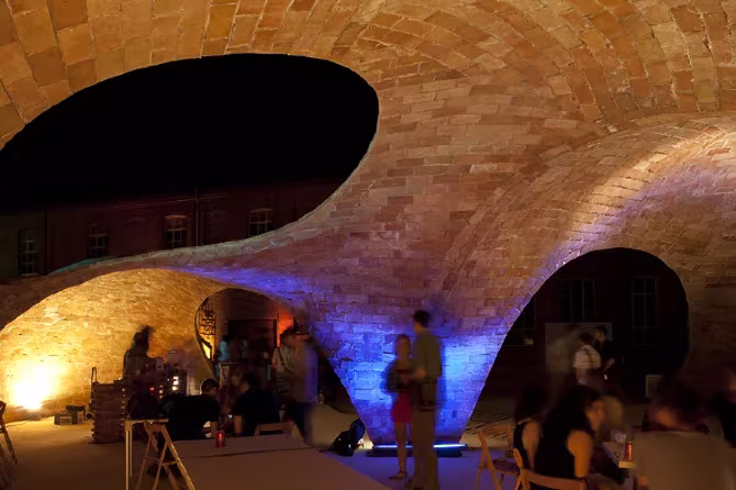 Pabellón Bricktopia de noche, con la bóveda de ladrillo iluminada en tonos cálidos y azules y gente debajo