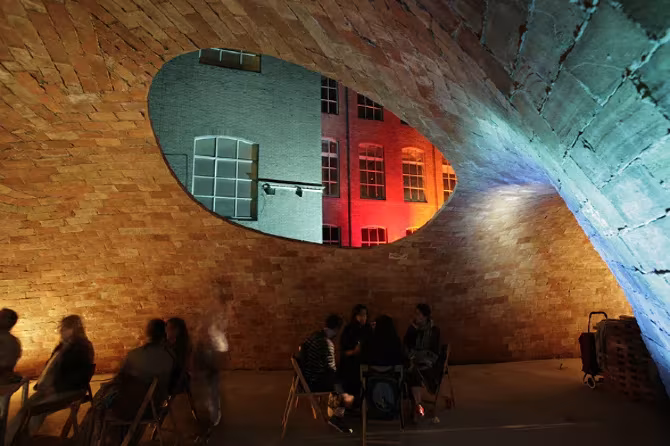 Abertura ovalada en la bóveda de Bricktopia que enmarca una fachada iluminada, con gente sentada debajo