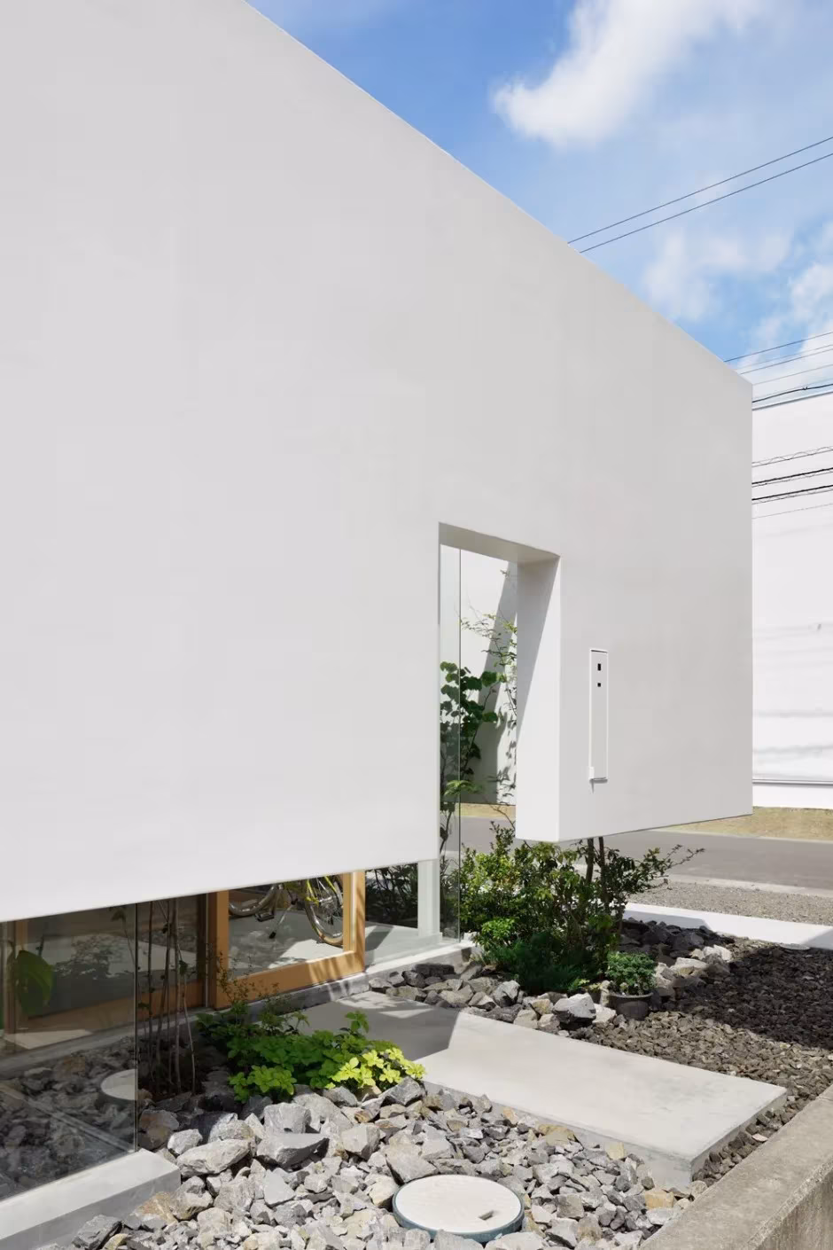Entrada de la Green Edge House: muro blanco con puerta estrecha y franja verde de piedras en la base