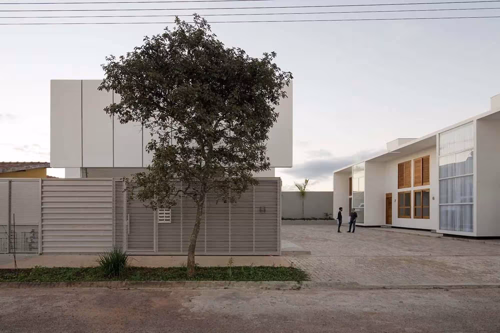Casas AV en Avaré al atardecer: volúmenes blancos tras un árbol y una valla, con dos personas en la acera