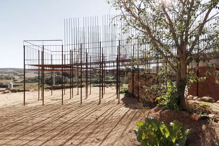 Estructura de varillas de acero oxidado que da sombra sobre un patio de tierra junto a un árbol en Seró
