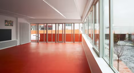 Sala alargada de suelo rojo junto a una fachada acristalada en las Escuelas Josephine Baker