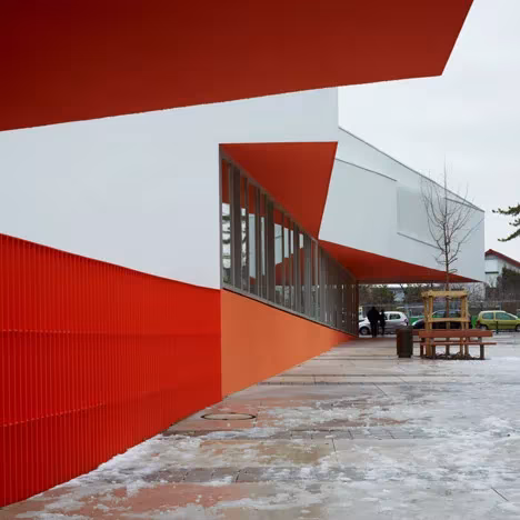 Porche cubierto de las Escuelas Josephine Baker con zócalo naranja, voladizo rojo y patio nevado
