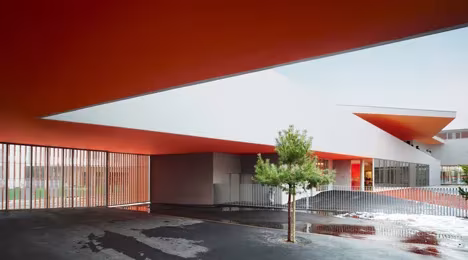 Voladizo naranja sobre el patio de las Escuelas Josephine Baker, con un pequeño pino y charcos de nieve
