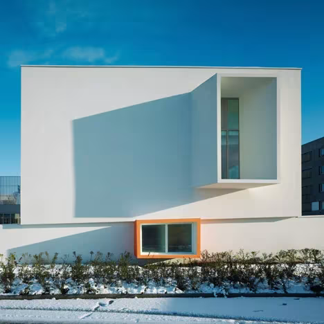 Volumen blanco de las Escuelas Josephine Baker con ventana saliente y hueco naranja sobre un seto nevado