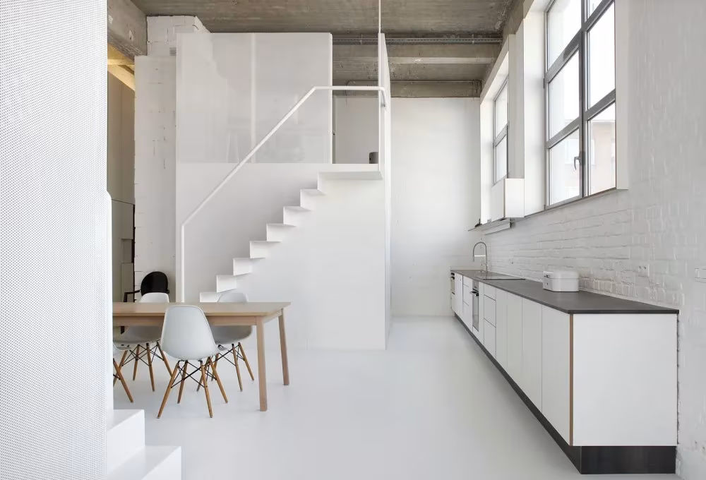 Cocina lineal blanca y escalera del Loft For junto a ventanales industriales y mesa con sillas