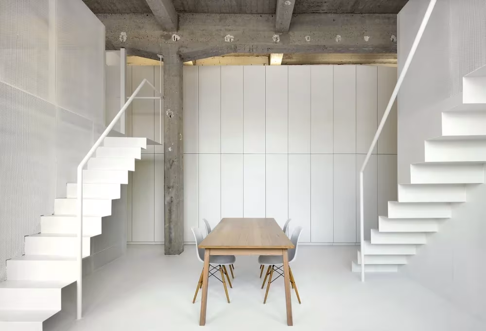 Dos escaleras blancas simétricas enmarcan el comedor del Loft For bajo vigas de hormigón