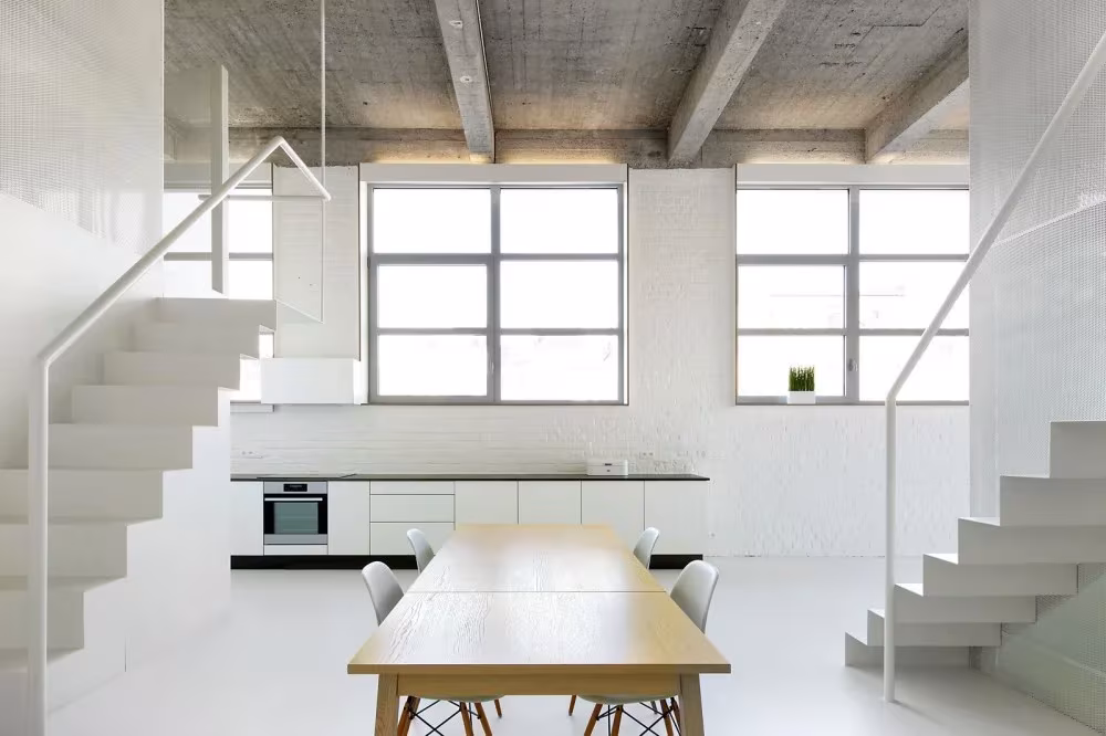 Mesa de madera larga ante los ventanales del Loft For, con escalera blanca y cocina al fondo