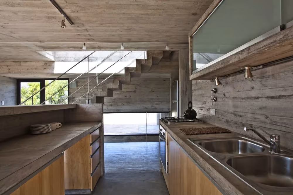 Encimera con fregadero de acero en una cocina de hormigón y madera con escalera al fondo