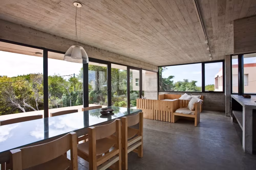 Salón comedor de hormigón con mesa de madera y grandes ventanales hacia la vegetación costera