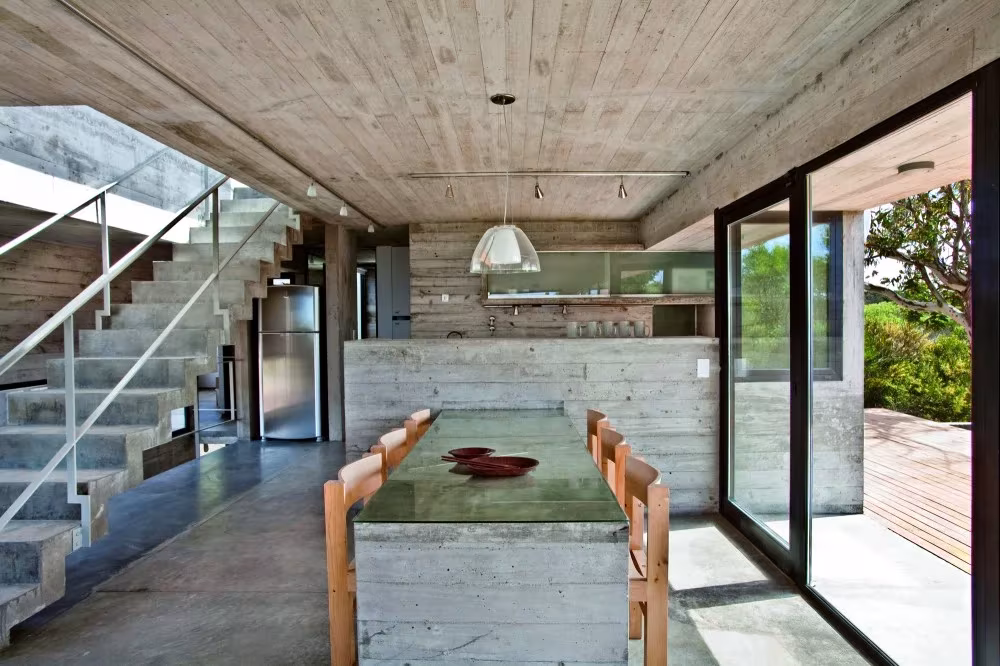 Comedor con mesa de hormigón, sillas de madera y escalera metálica en la casa de BAK Arquitectos