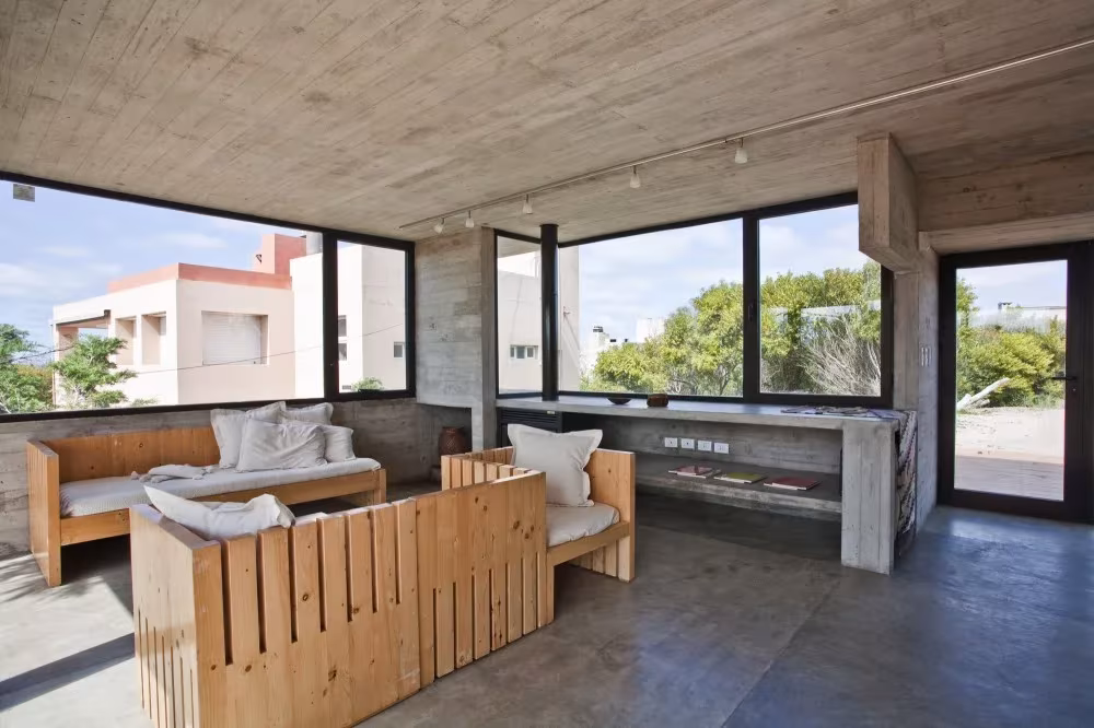 Salón de hormigón con sofás de listones de madera y ventanales hacia la vegetación de la costa