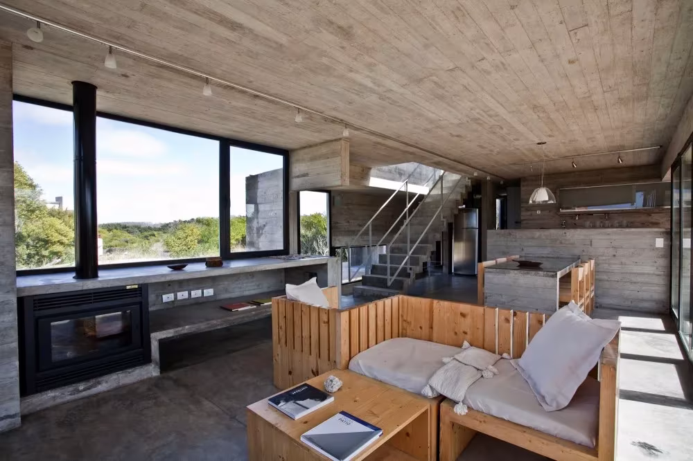 Salón con chimenea empotrada, sofá de madera con cojines blancos y escalera al fondo en una casa de hormigón