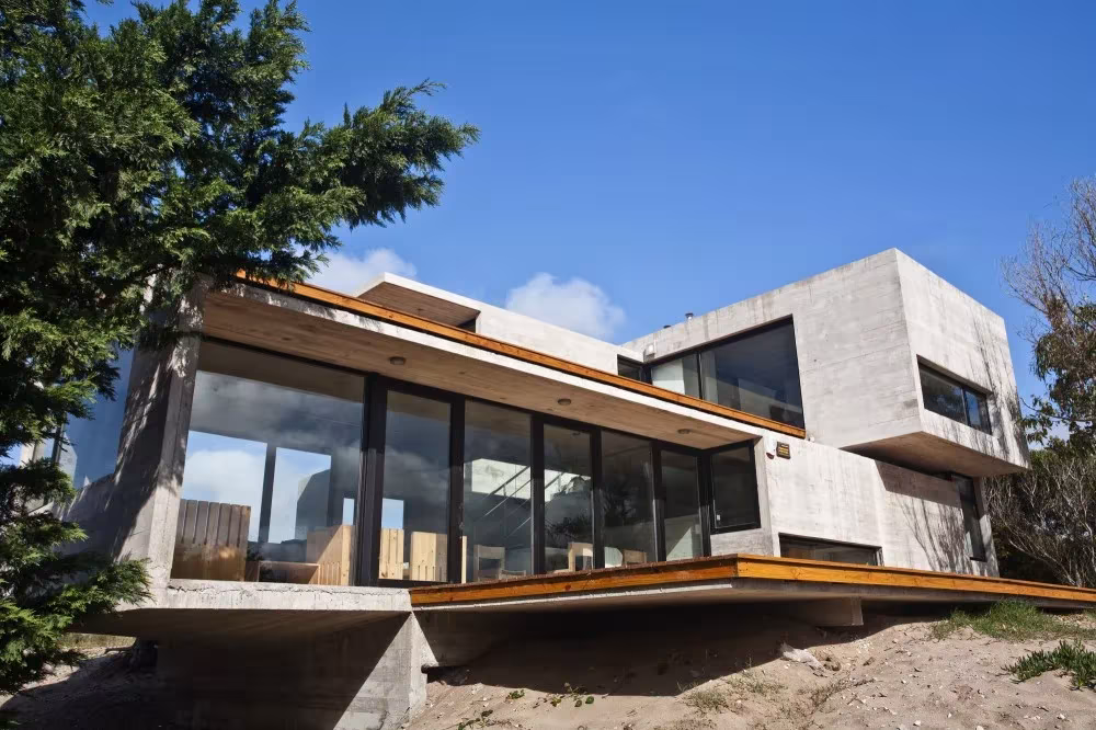 Volúmenes de hormigón y vidrio de la Casa en la Playa entre árboles, con terraza en voladizo
