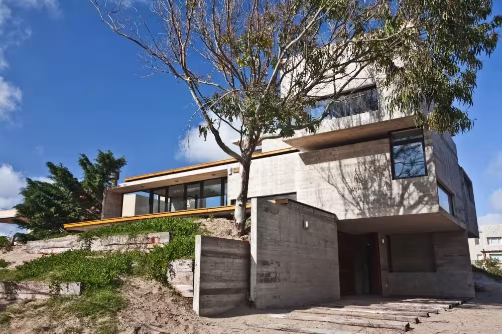 Casa de hormigón encofrado con tablas semienterrada en la duna junto a un árbol, en la costa bonaerense
