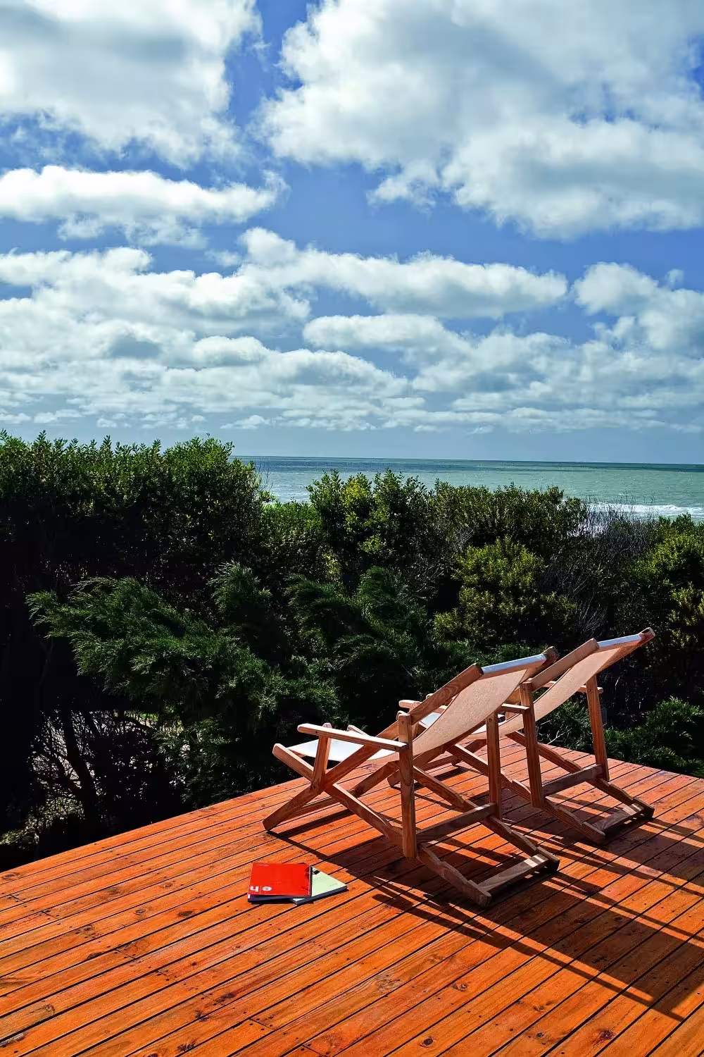 Dos tumbonas de madera en una terraza con vistas al mar entre la vegetación y cielo con nubes