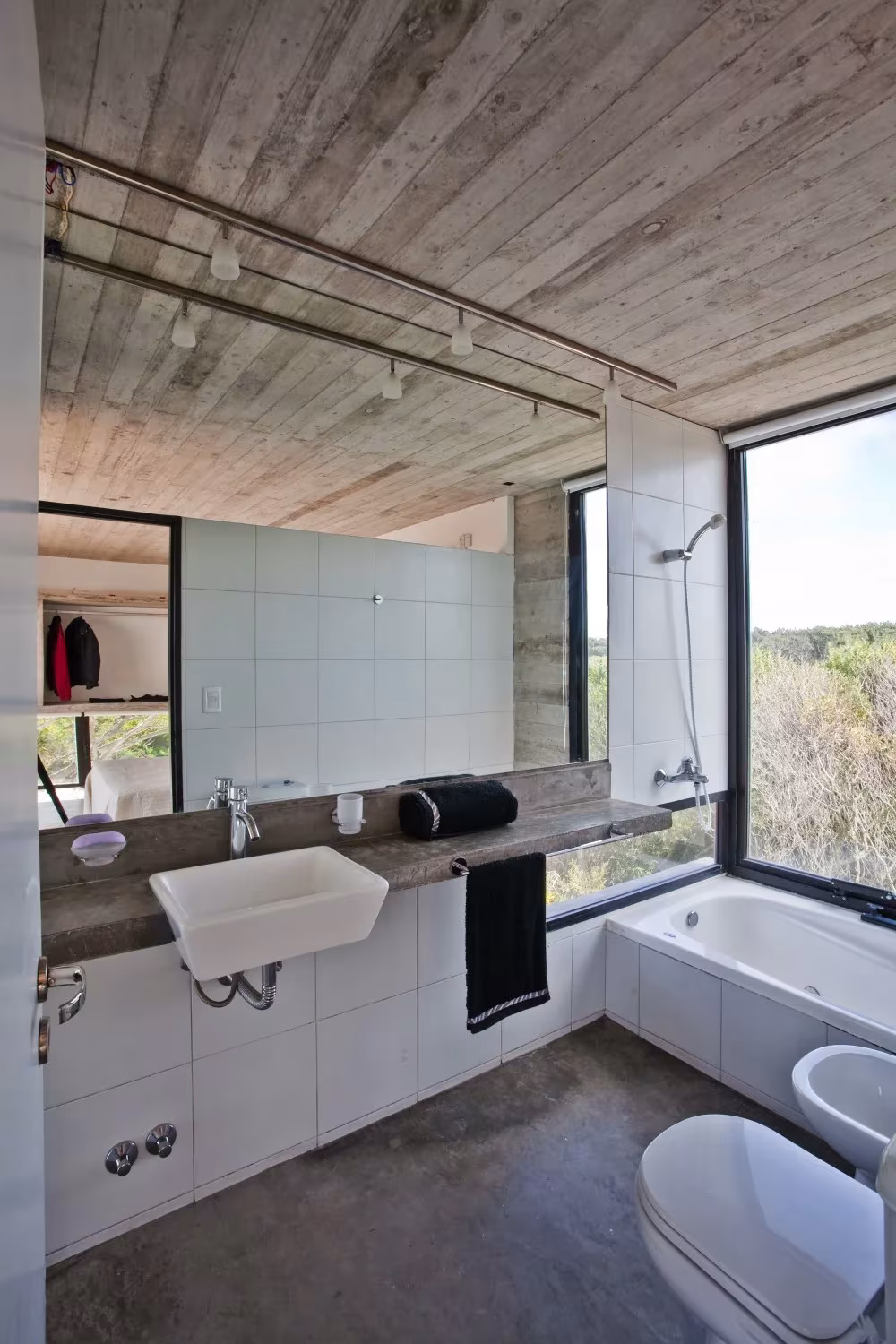Baño con lavabo blanco, bañera junto a una ventana y techo de hormigón con huella de tabla