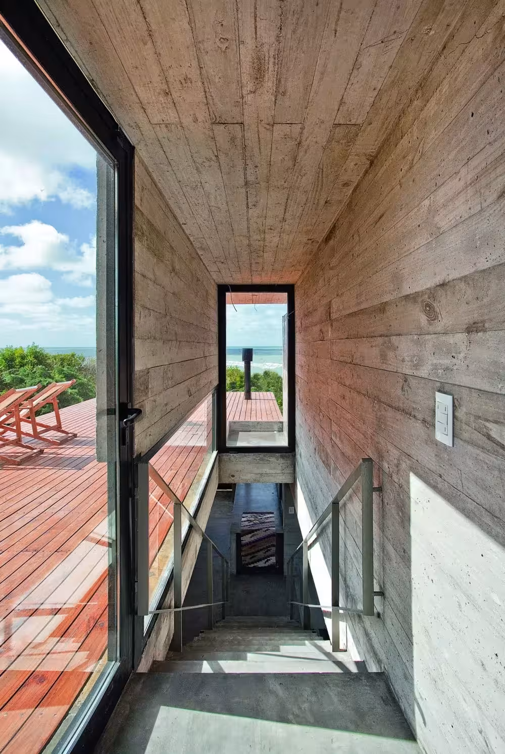 Pasillo estrecho de hormigón con hueco de escalera y puerta abierta a una terraza de madera