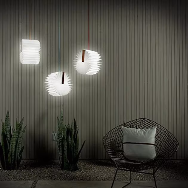 Lámparas Lumio colgadas en un patio nocturno junto a una silla de rejilla metálica y plantas