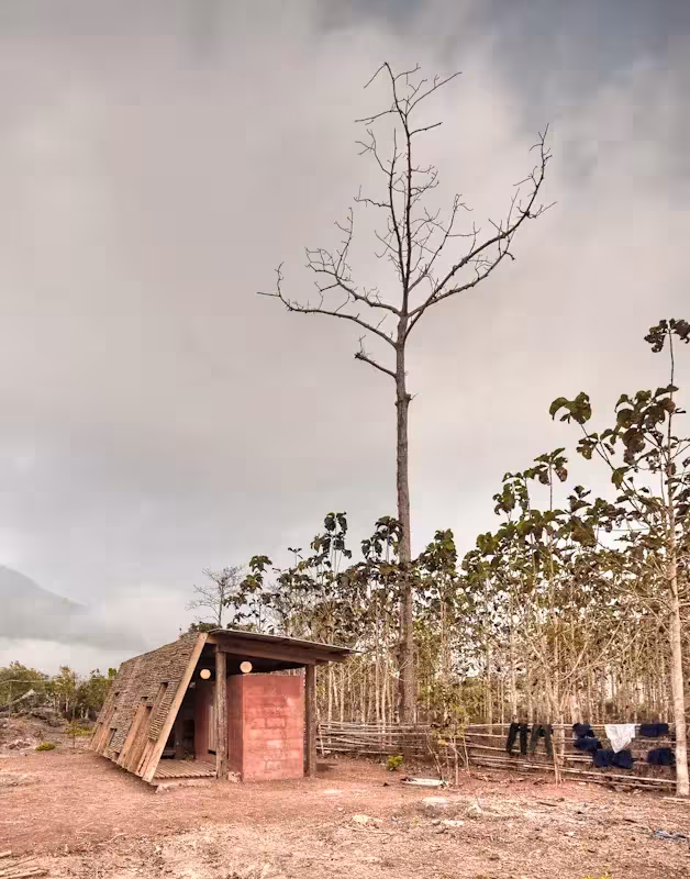Pequeña casa de baños de bambú en un terreno árido junto a un árbol seco bajo un cielo nublado