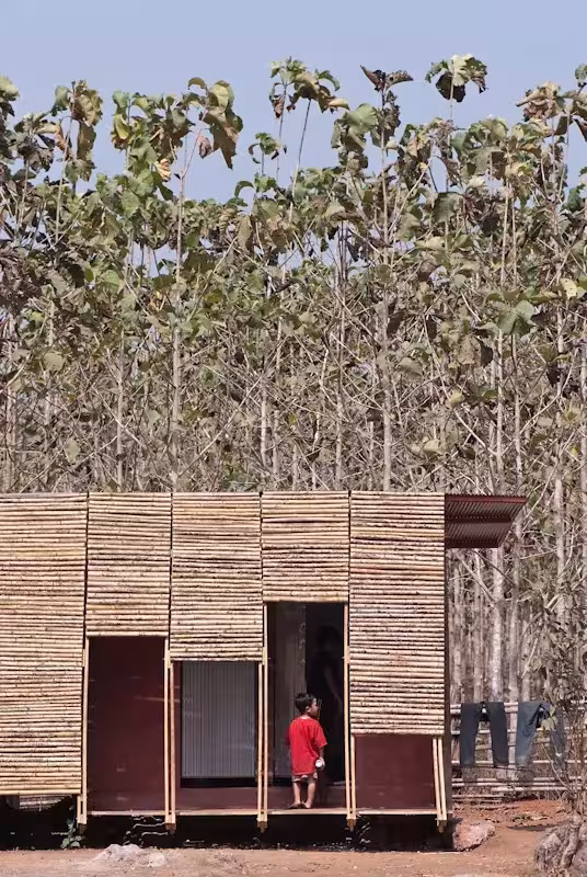 Fachada de cañas de bambú de la casa de baños con un niño en la puerta y árboles de teca detrás