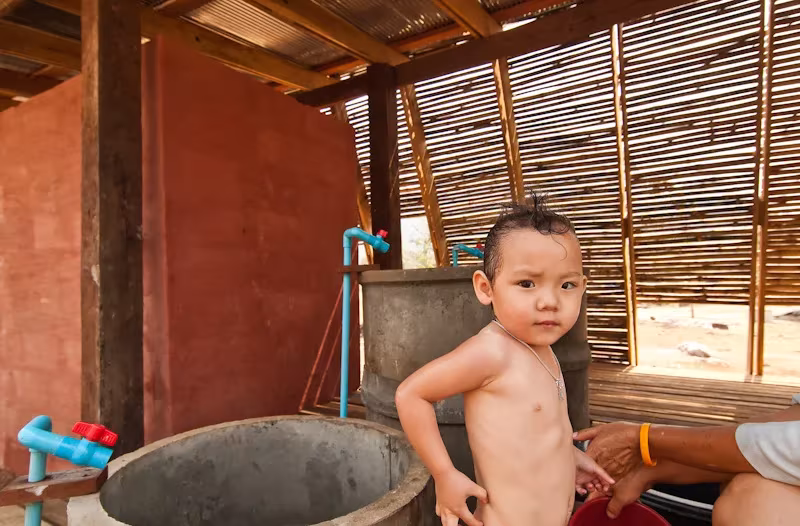 Niño pequeño junto a un depósito de agua de hormigón en la casa de baños del orfanato