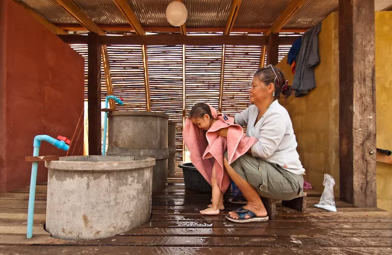 Mujer secando con una toalla a una niña junto a los depósitos de agua de la casa de baños