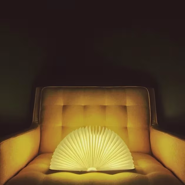 Lámpara Lumio abierta en abanico iluminando un sillón capitoné en la oscuridad