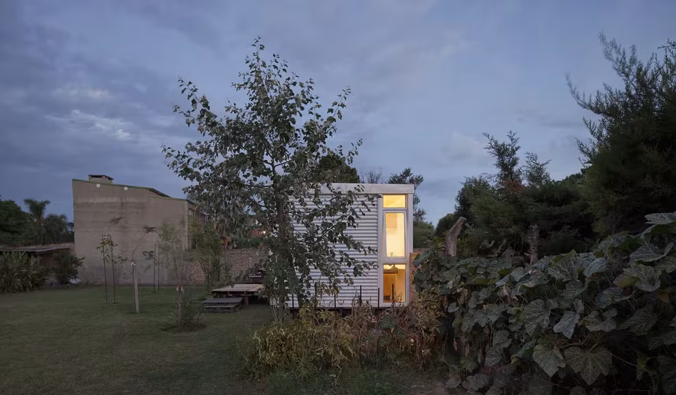 Mia House al anochecer con una ventana iluminada entre árboles y vegetación