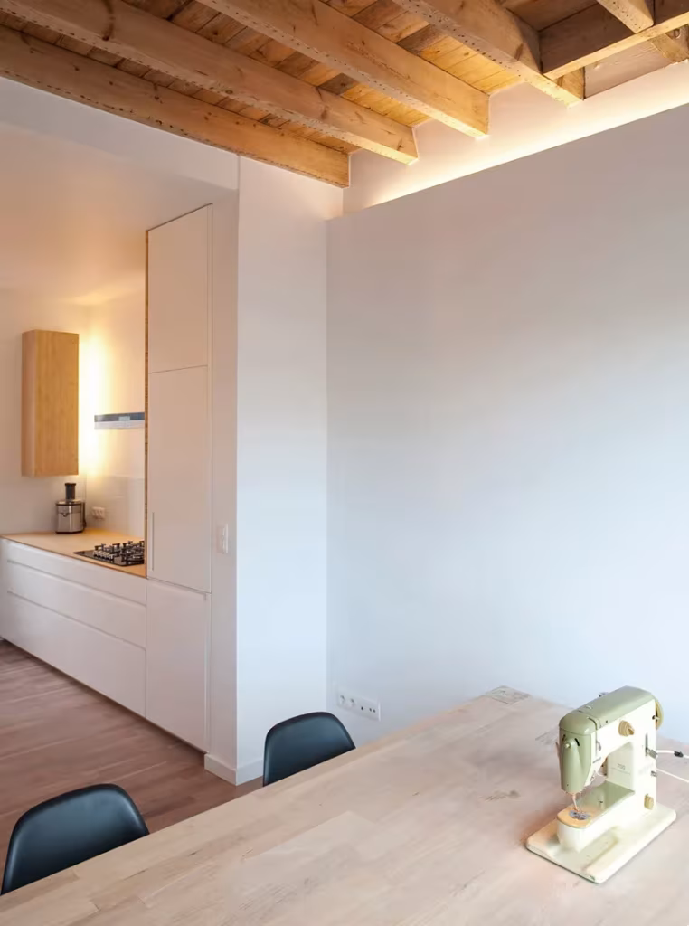 Cocina blanca bajo vigas de madera vista con mesa de madera y sillas negras en la Casa KCV