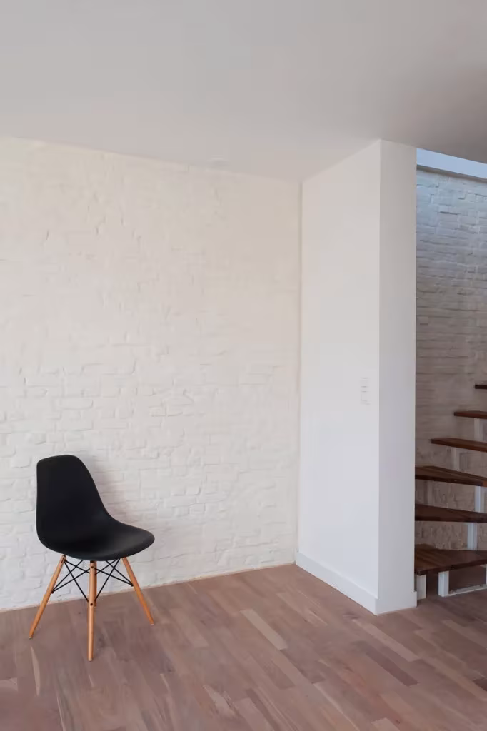 Silla negra junto a un muro de ladrillo pintado de blanco y suelo de madera en la Casa KCV