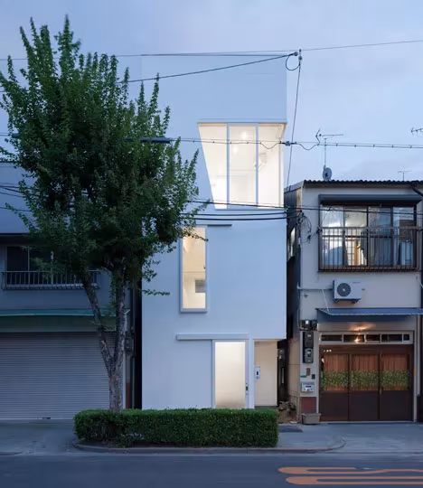 Casa Tamatsu al atardecer, estrecha y blanca entre edificios vecinos, con ventanas iluminadas