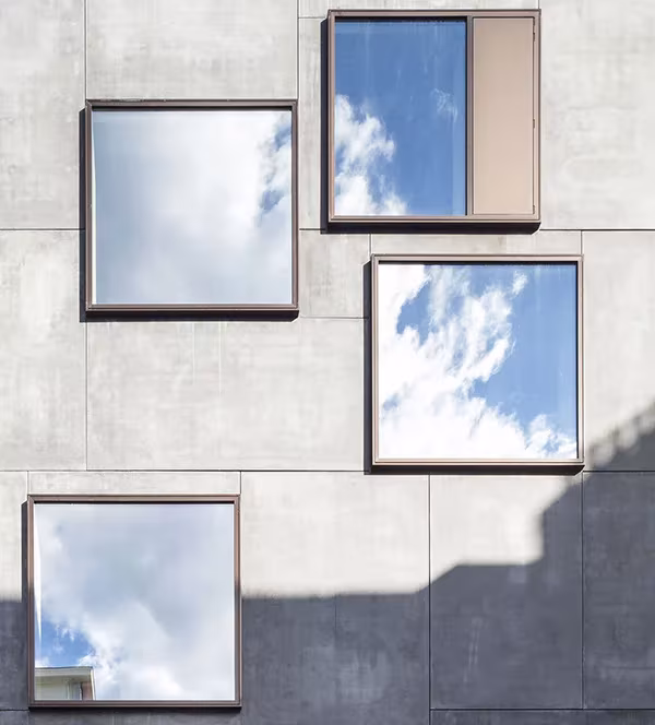 Fachada de hormigón del Little Willy en Bruselas con ventanas cuadradas de marco marrón que reflejan el cielo