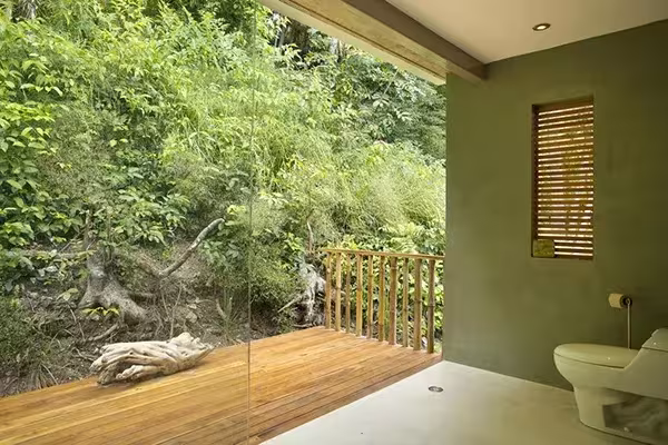 Baño de la Casa Flotante abierto a la vegetación tropical, con suelo de madera, paredes verdes y barandilla