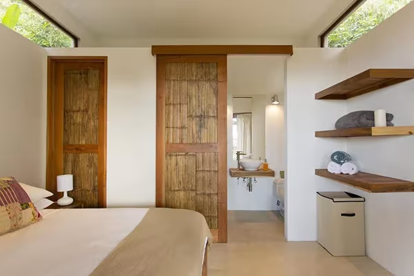 Dormitorio de la Casa Flotante con paredes blancas, puertas de bambú, baldas de madera y baño al fondo