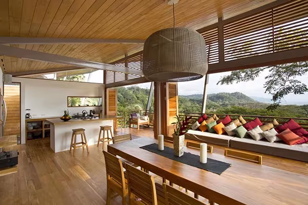 Salón abierto de la Casa Flotante con cocina, mesa larga, banco con cojines de colores y lamas de madera