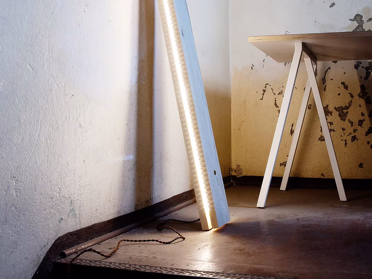 Lámpara lineal de madera apoyada contra la pared junto a una mesa de caballetes blancos