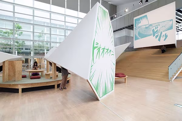 Paneles blancos con ilustraciones verdes y gradas de madera en la exposición del YCAM bajo la fachada de vidrio