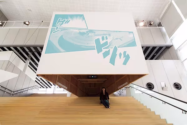Gran panel con dibujo de olas sobre la escalera de madera del YCAM, con una persona sentada debajo