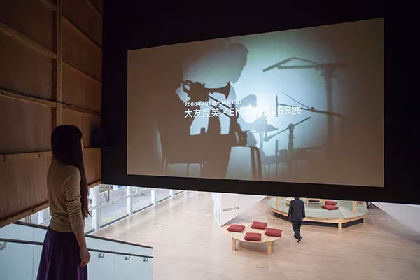 Proyección en gran pantalla dentro de la exposición del YCAM, con visitantes y cojines rojos en la sala