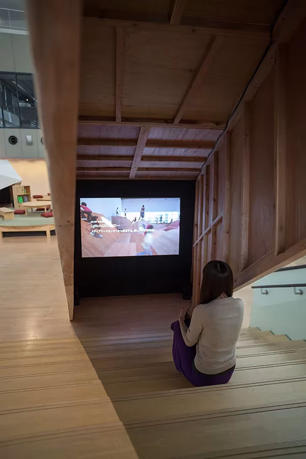 Persona sentada en la grada de madera del YCAM viendo un vídeo en una pequeña sala de proyección