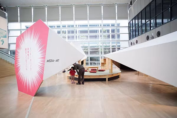 Panel rosa inclinado en la exposición del YCAM con visitantes junto a una plataforma de madera al fondo