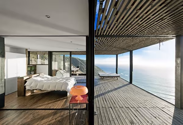 Dormitorio acristalado junto a una terraza de madera con celosía y vistas al océano en la Casa Till